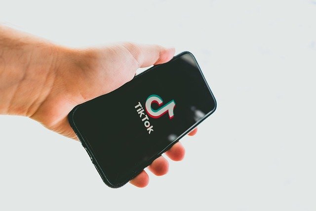 A hand holding a smartphone displaying the TikTok logo on a black screen, against a light background. The image conveys a modern, tech-savvy tone.