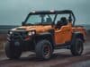 A Can Am Defender UTV in orange and black color navigating a dirt road with trees lining the path.