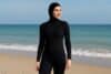 A woman in a full coverage black wetsuit stands on the beach, ready for a day of water activities.