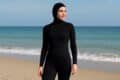 A woman in a full coverage black wetsuit stands on the beach, ready for a day of water activities.