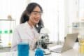 A woman in a lab coat examines a microscope, representing her role as an online chemistry tutor.