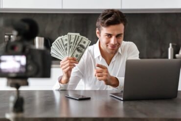 A man sits in front of a laptop, holding money, representing YouTube funds and online earnings.