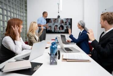 Business professionals in a meeting room using a laptop and a 360 conference camera for a video call.