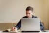 A man sitting at a table, focused on his laptop, with a pen in hand, ready to take notes or brainstorm ideas.