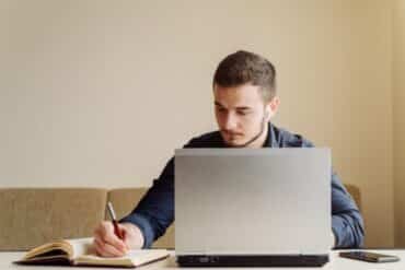 A man sitting at a table, focused on his laptop, with a pen in hand, ready to take notes or brainstorm ideas.
