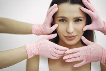 A woman wearing gloves is gently touching her face with a thoughtful expression.