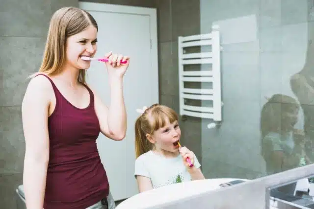 A woman and her young daughter happily brush their teeth together in a colorful bathroom, sharing a joyful routine.