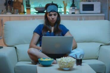 A woman relaxes on a couch with a laptop and popcorn, enjoying a cozy moment while watching 'Goonierne 2'.