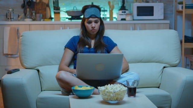 A woman relaxes on a couch with a laptop and popcorn, enjoying a cozy moment while watching 'Goonierne 2'.