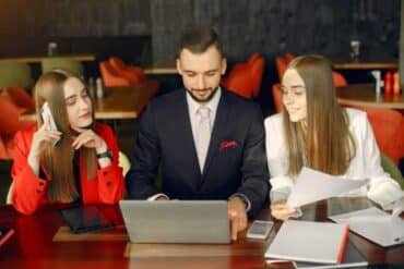 Three professionals in suits gather around a table, engaged in conversation with a laptop between them.