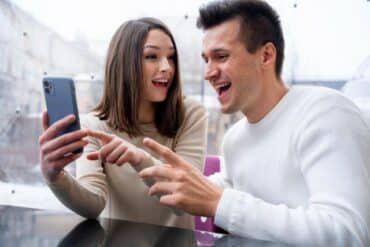 A man and woman laugh together while looking at a cell phone, enjoying content from Just For Fans videos.