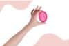 A hand holding a pink menstrual disc against a matching pink background.