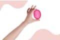 A hand holding a pink menstrual disc against a matching pink background.