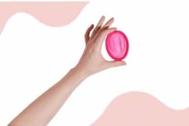 A hand holding a pink menstrual disc against a matching pink background.