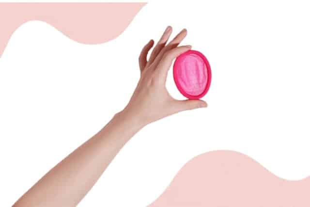 A hand holding a pink menstrual disc against a matching pink background.