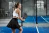 A woman in a white tank top and black skirt plays tennis at NLPadel, focused on hitting the ball.