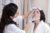 A woman receives a facial examination from a doctor in a bright, clinical setting.