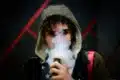 A man wearing a hoodie is smoking an electronic cigarette, specifically a Foger vape.