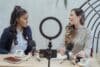Two women sit at a table with microphones and coffee, engaged in conversation and sharing ideas.