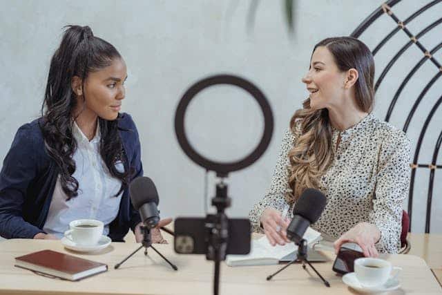 Two women sit at a table with microphones and coffee, engaged in conversation and sharing ideas.