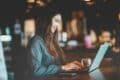 A woman sits at a table in a cafe, smiling while typing on her laptop as she manages Multi-Channel Marketing Automation, with a glass of water beside her.