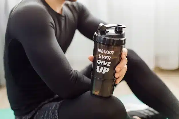A man sits on a yoga mat, holding a shaker bottle, ready for his workout or post-exercise refreshment.