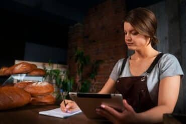 A woman in an apron writes on a tablet, planning her bakery business with a focused expression.