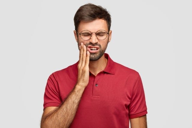 A man in a red polo shirt and glasses grimaces, pressing his hand to his cheek, suggesting dental pain or discomfort. Gray background conveys a neutral mood.