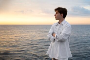 A young man in a white shirt stands at the ocean's edge, reflecting on therapeutic hobbies for a stress-free mind.