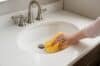 A person cleaning an undermount bathroom sink with a yellow cloth, focusing on removing dirt and grime.