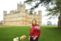 A woman in a red sweater and jeans stands with two dogs in front of Abbey Castle in London.