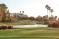 A scenic view of the David L Baker golf course featuring lush green fairways, a calm water hazard, tall palm trees, and a clubhouse in the background under a clear sky.