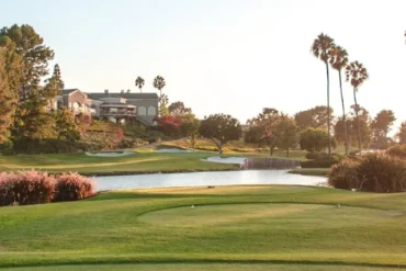 A scenic view of the David L Baker golf course featuring lush green fairways, a calm water hazard, tall palm trees, and a clubhouse in the background under a clear sky.