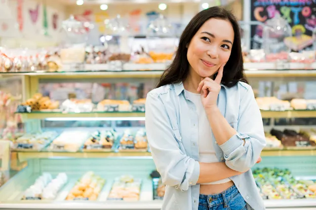 The image shows a young woman standing in front of a pastry display, looking thoughtfully at the treats on display. She has long, dark hair, and is wearing a light blue shirt over a white top, with her arms crossed and her finger placed on her chin. The background features a variety of baked goods neatly arranged in the store's display case, giving the setting a cozy, inviting atmosphere.
