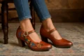 A close-up of a pair of L’Artiste women’s leather shoes with chunky heels, one decorated with vibrant floral embroidery and a gold buckle strap, worn with blue jeans, resting on a patterned antique rug.