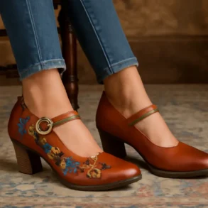 A close-up of a pair of L’Artiste women’s leather shoes with chunky heels, one decorated with vibrant floral embroidery and a gold buckle strap, worn with blue jeans, resting on a patterned antique rug.