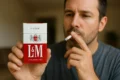 A man holds a red-and-white pack of L&M cigarettes in one hand while smoking a cigarette with the other, looking contemplative.