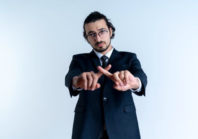 Business man in black suit and glasses looking to the front with serious face crossing index fingers making defense gesture standing over white wall.