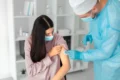 A healthcare professional in protective gear administers an onsite flu vaccination to a patient, as they receive the shot in a clean, modern medical setting. The patient, wearing a face mask, looks down at their arm while the caregiver gently applies a cotton ball to the injection site.