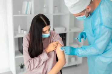 A healthcare professional in protective gear administers an onsite flu vaccination to a patient, as they receive the shot in a clean, modern medical setting. The patient, wearing a face mask, looks down at their arm while the caregiver gently applies a cotton ball to the injection site.