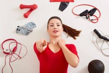 A woman stands among various fitness equipment, including weights and mats, in a well-equipped gym setting.