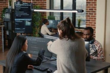 Software programer pointing pencil at source code on computer screen explaining algorithm to coworker standing next to desk. programmers discussing online cloud computing with team.