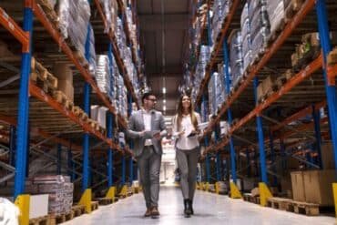 Two people walking side by side down a wide aisle in a busy warehouse filled with shelves and boxes.