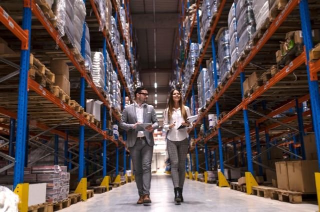 Two people walking side by side down a wide aisle in a busy warehouse filled with shelves and boxes.