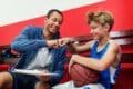 A man and a boy sit together in a gym, holding a basketball and enjoying their time on the court.