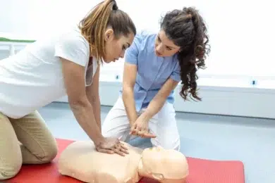 Two women performing CPR on a mannequin during a first aid training session. One woman, kneeling beside the mannequin, is giving chest compressions, while the other, standing next to her, is offering guidance on the proper technique. The setting appears to be a medical or training facility with a clean and bright environment.