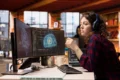 A woman with headphones sits at a desk, focused on two monitors, providing IT support with AI assistance.