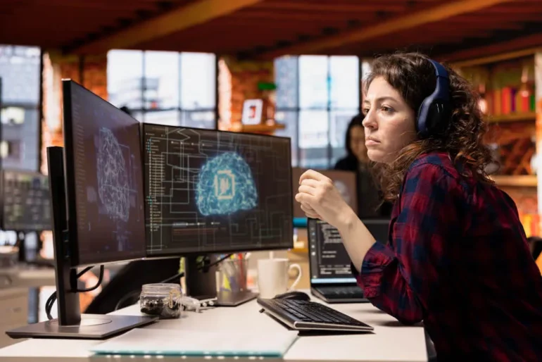 A woman with headphones sits at a desk, focused on two monitors, providing IT support with AI assistance.