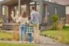 A family of four stands together in front of their house, smiling and posing for the camera.