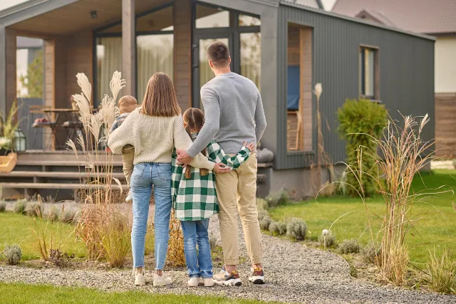 A family of four stands together in front of their house, smiling and posing for the camera.